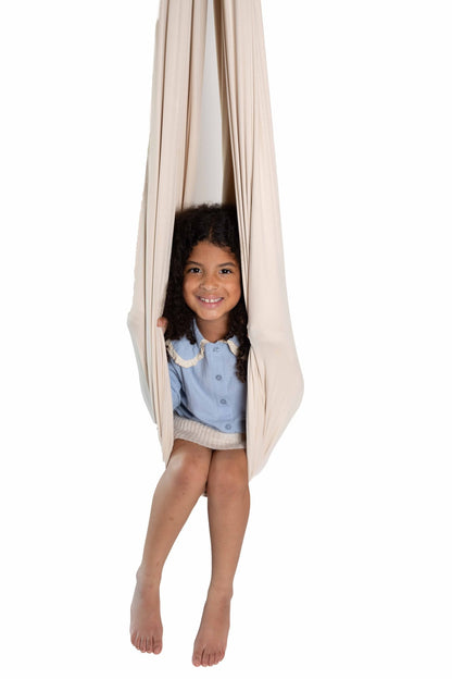 A smiling child sitting in a cream sensory swing against a white background